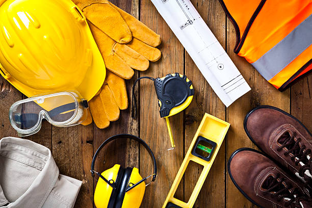 Personal protective workwear and blueprint with some measuring instruments shot directly from above on rustic wood background. The protective workwear includes hard hat, gloves, earmuff, goggles, steel toe shoes, and safety vest. The composition also includes a tape measure and bubble level and a construction blueprint, all items used by construction worker or engineer. Predominant colors: yellow and brown.